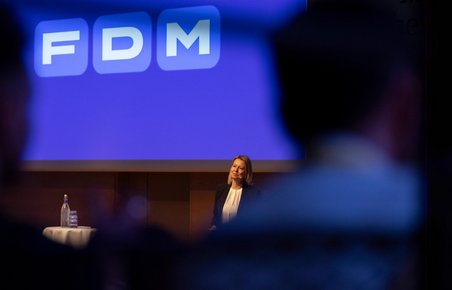 FDMs administrerende direktør, Stina Glavind, holder oplæg foran blå skærm med FDM-logo. Silhouetter af mennesker i forgrunden.